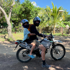 Two people wearing helmets ride a motorcycle outdoors on a sunny day, surrounded by trees and plants. One of them carries the Packable Bullet Ruck - Ripstop ROBIC®, a lightweight travel backpack perfect for their adventure.