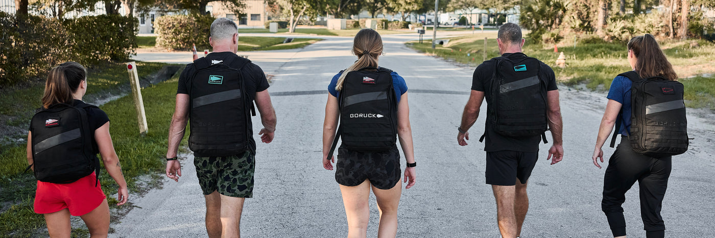 Five people carrying the Ruck Bundle backpacks walk down a sunlit street, viewed from behind.