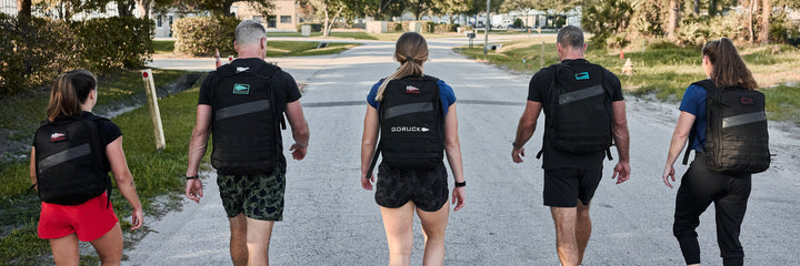 Five people carrying the Ruck Bundle backpacks walk down a sunlit street, viewed from behind.