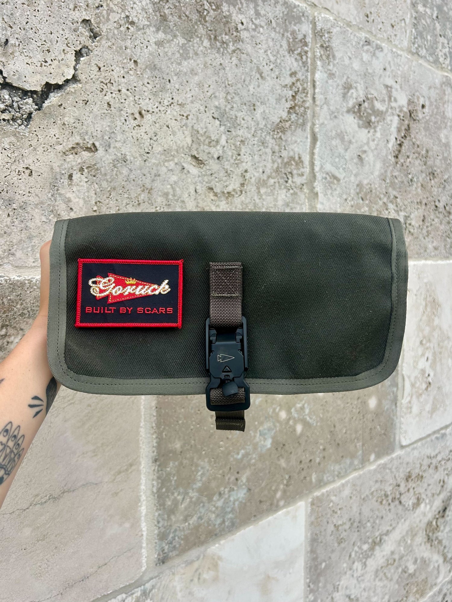 A hand holds the Scars Wire Dopp, a green waxed twill pouch with a red 