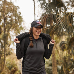 A smiling woman in outdoor gear and a Performance TAC Hat - TOUGHDRY stands in a forest, holding her jacket over her shoulders.