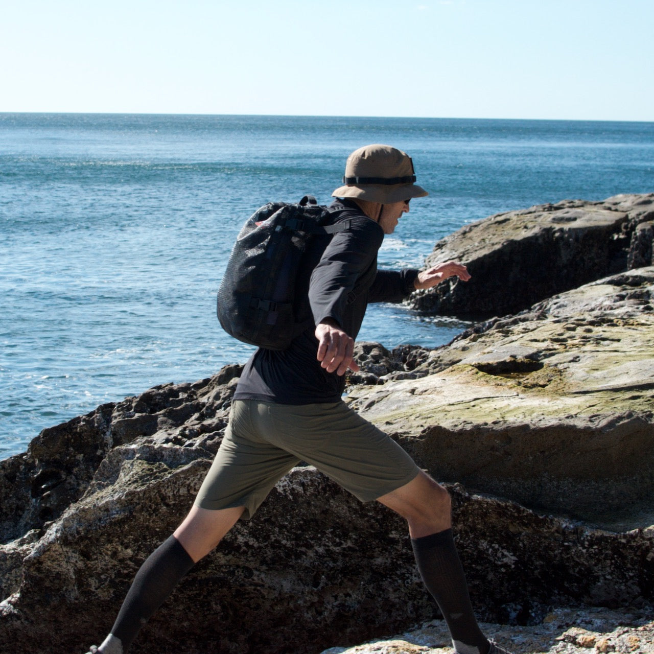 Wearing the Boonie Hat - Tactical - ToughDry and a backpack, a person steps across the rocky ocean shore on a sunny day.