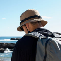A person wearing a tan Boonie Hat - Tactical - ToughDry and backpack stands by the ocean on a sunny day, facing away. The military-style hat is made from durable ToughDry fabric, ideal for outdoor adventures.