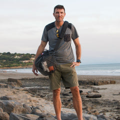 A man stands on a rocky beach with the Mesh Gym Bag slung over his shoulder, dressed in shorts and a t-shirt, with the ocean and hills behind him.