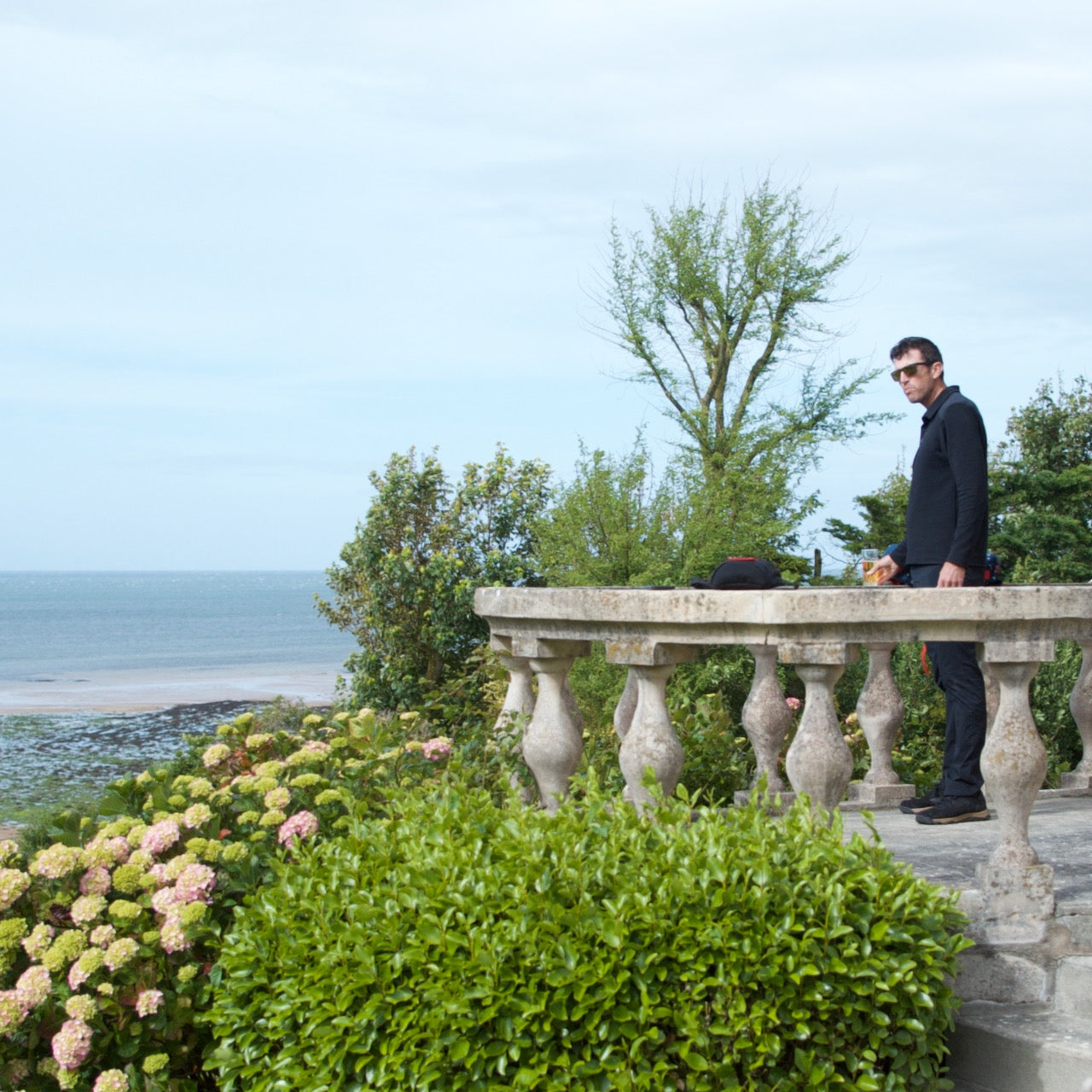 A man in sunglasses stands on a stone terrace overlooking the sea, surrounded by lush greenery and flowers, wearing the Men's Rucking Shirt - Merino Wool.