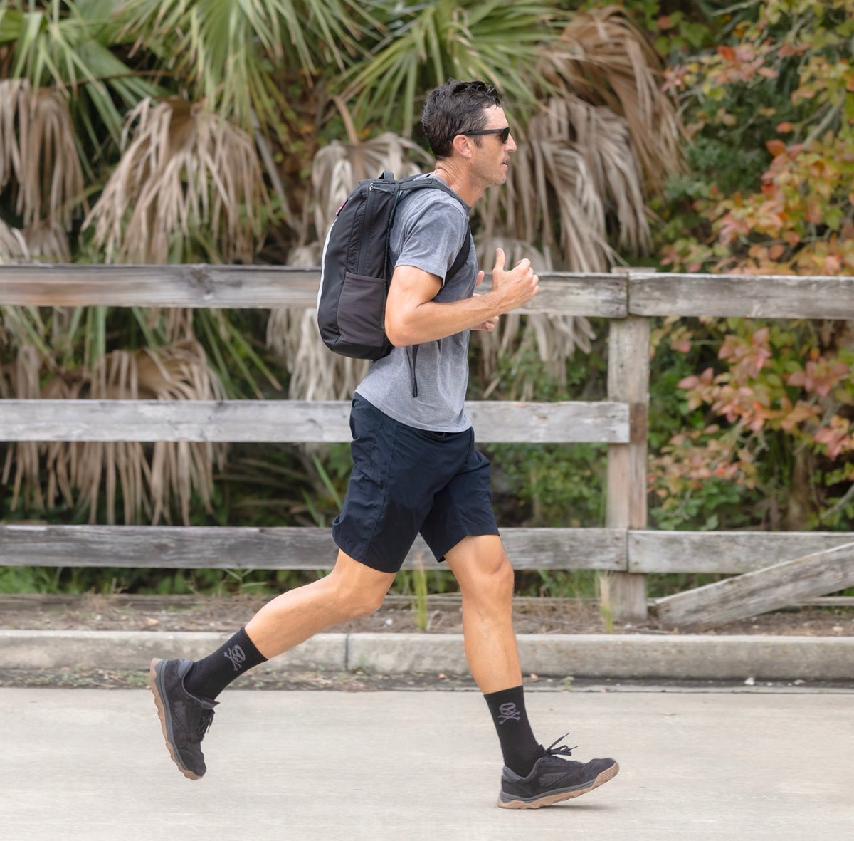 A man jogs on a sidewalk wearing a gray shirt, black shorts, black shoes, and carrying the Basic Rucker backpack.