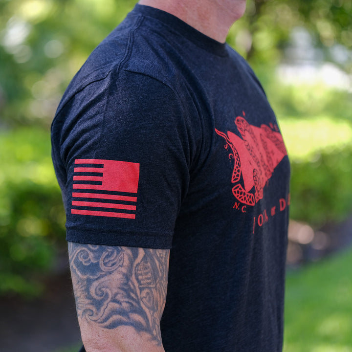 A man outdoors wears the Join or Die Tee - Tri-Blend, featuring a red flag and snake design from Ben Franklin—a tribute to colonial unity—while displaying his tattooed arm.