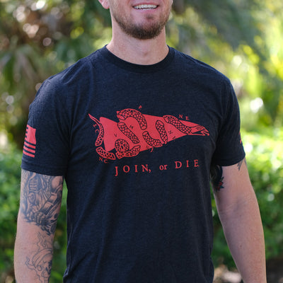 A smiling man stands outdoors in the Join or Die Tee - Tri-Blend, featuring a red segmented snake graphic inspired by Ben Franklin's iconic colonial unity message.