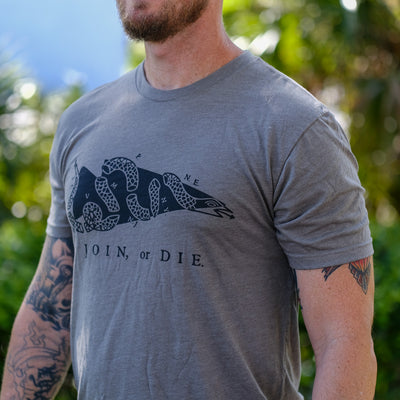 A man stands outdoors wearing the Join or Die Tee - Tri-Blend, featuring Ben Franklin’s iconic segmented snake design symbolizing colonial unity.