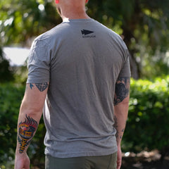 Back view of a man wearing a grey GORUCK t-shirt outdoors with visible arm tattoos and greenery background