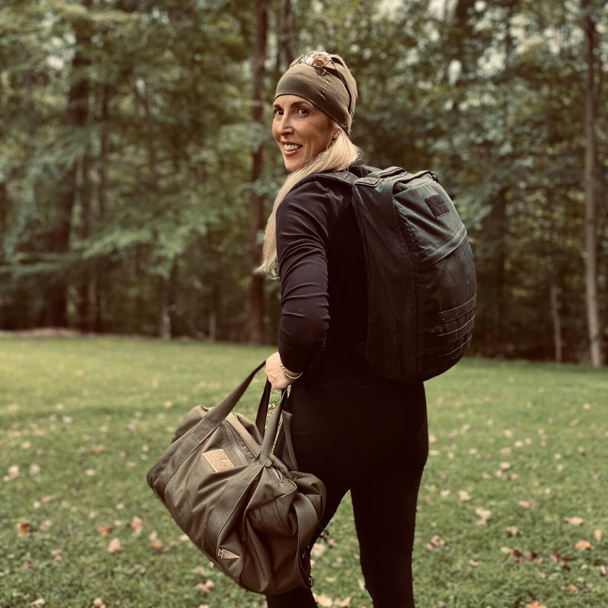 Woman in athletic wear, carrying a backpack and duffel bag, stands outdoors in a grassy, wooded area.