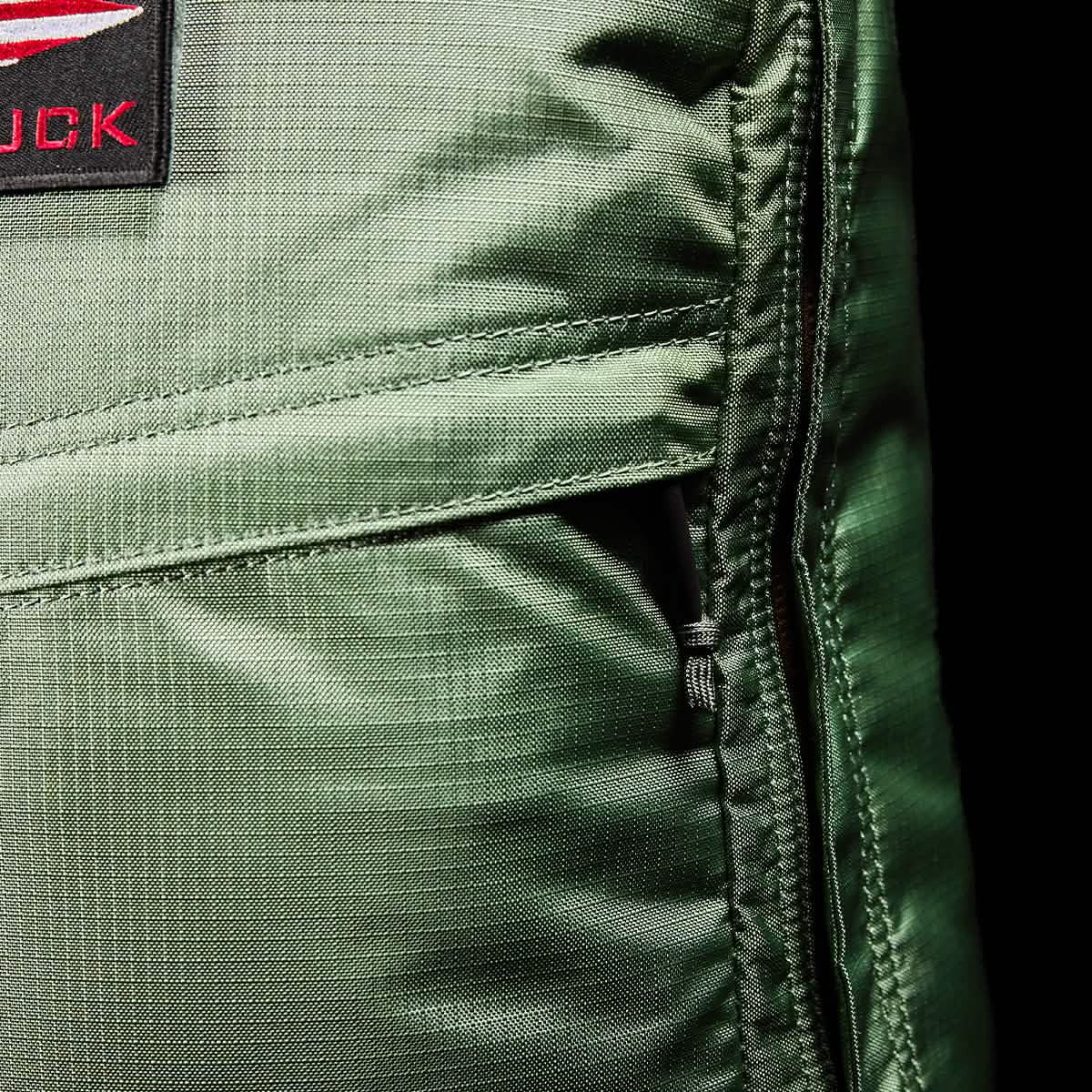 Close-up of the GR1 USA - Ripstop ROBIC® rucksack by GORUCK, showcasing its lightweight green design with a red and black patch that highlights visible stitching and a right-side zipper. The sheen of the water-resistant fabric is evident under the lighting.