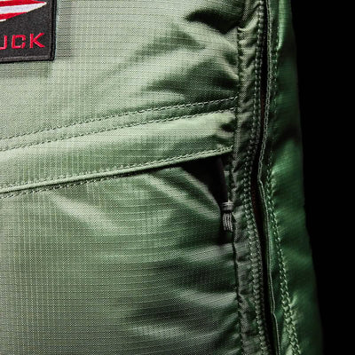 Close-up of the GR1 USA - Ripstop ROBIC® rucksack by GORUCK, showcasing its lightweight green design with a red and black patch that highlights visible stitching and a right-side zipper. The sheen of the water-resistant fabric is evident under the lighting.