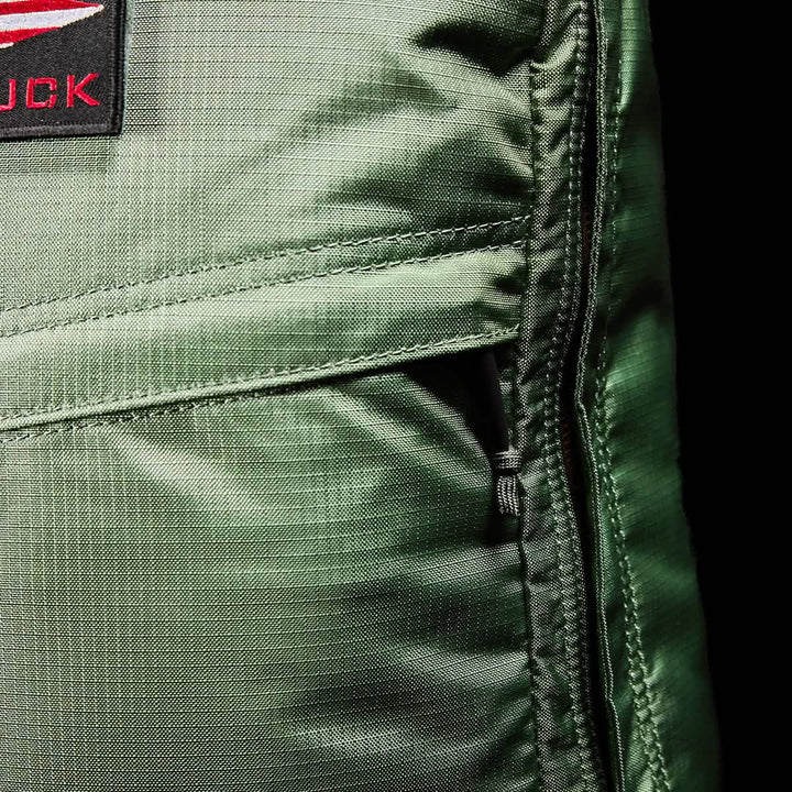 Close-up of the GR1 USA - Ripstop ROBIC® rucksack by GORUCK, showcasing its lightweight green design with a red and black patch that highlights visible stitching and a right-side zipper. The sheen of the water-resistant fabric is evident under the lighting.