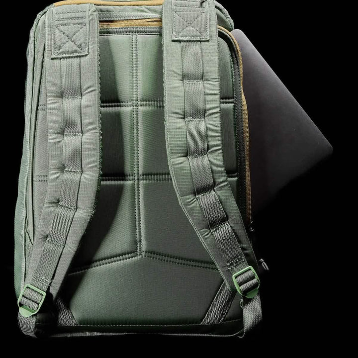 Against a black background stands the GR1 USA - Ripstop ROBIC® rucksack by GORUCK, showcasing its lightweight design in green. The bag features quilted straps and is partially open to reveal the edge of a black laptop inside. Its textured, water-resistant fabric highlights its durability.