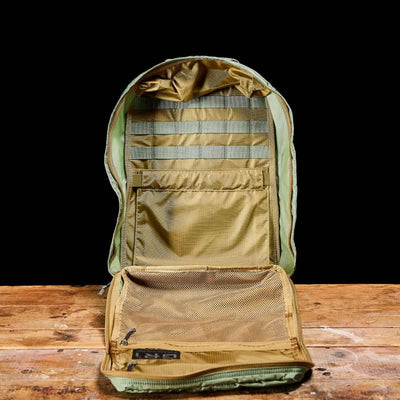 The GORUCK GR1 USA - Ripstop ROBIC® rucksack, in olive green, rests on a wooden surface against a black background. Its lightweight interior includes multiple compartments with mesh pockets and horizontal strap organizers, offering ample storage space. Designed to be water-resistant, it's perfect for any adventure.