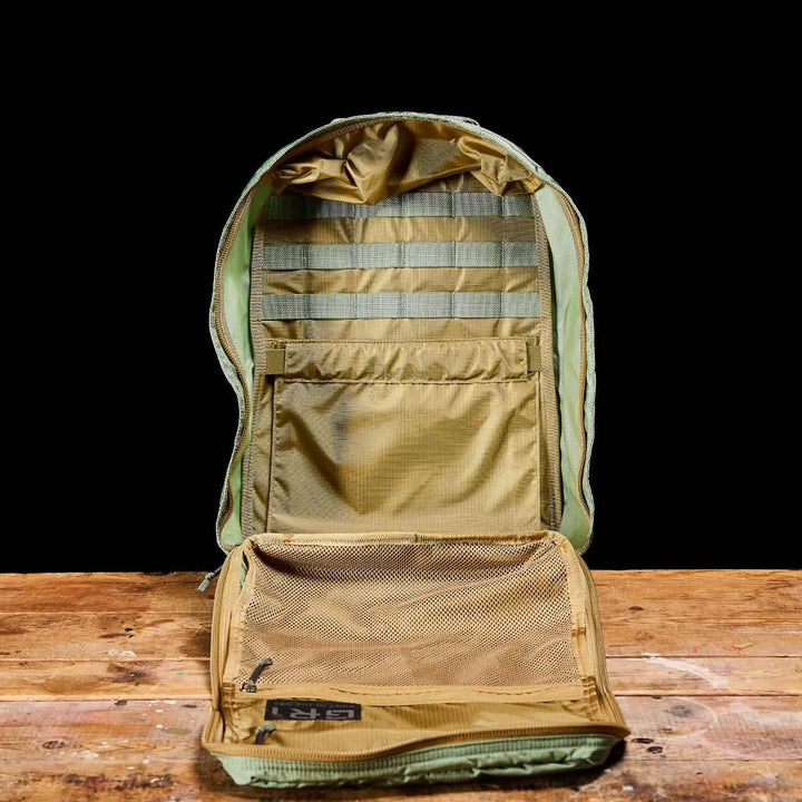 The GORUCK GR1 USA - Ripstop ROBIC® rucksack, in olive green, rests on a wooden surface against a black background. Its lightweight interior includes multiple compartments with mesh pockets and horizontal strap organizers, offering ample storage space. Designed to be water-resistant, it's perfect for any adventure.