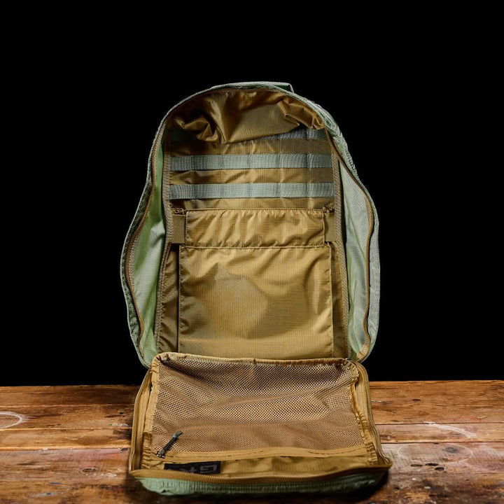 The GR1 USA - Ripstop ROBIC® by GORUCK, an ultra-lightweight rucksack in green and brown, is open to reveal its multiple compartments and a mesh pocket inside. This water-resistant bag is elegantly displayed against a black background on a wooden surface.