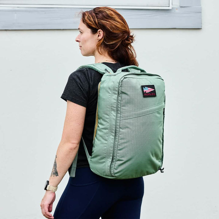 A person with reddish-brown hair is facing sideways, sporting a black T-shirt and blue pants, while carrying an ultra-lightweight green GORUCK GR1 USA - Ripstop ROBIC® rucksack. They have a tattoo on their right forearm and are standing against an off-white wall.