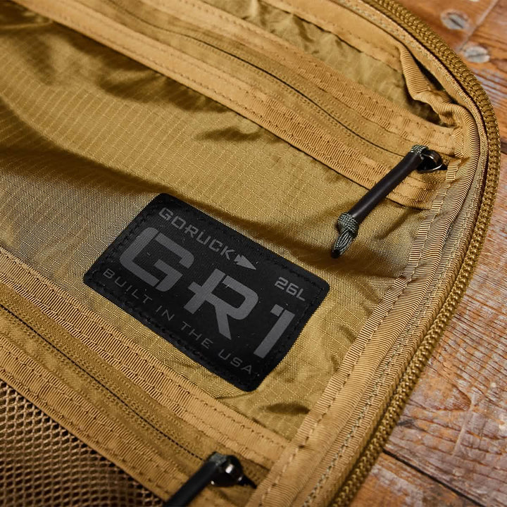 Close-up of an open GORUCK rucksack showcasing a label that reads GR1 USA - Ripstop ROBIC® Built in the USA. The ultra-lightweight tan interior features mesh pockets and sturdy, water-resistant zippers, all set on a wooden surface.