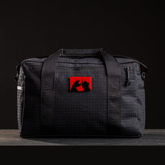 Black rucksack with dual handles and red patch showing samurai silhouettes, set on wooden surface