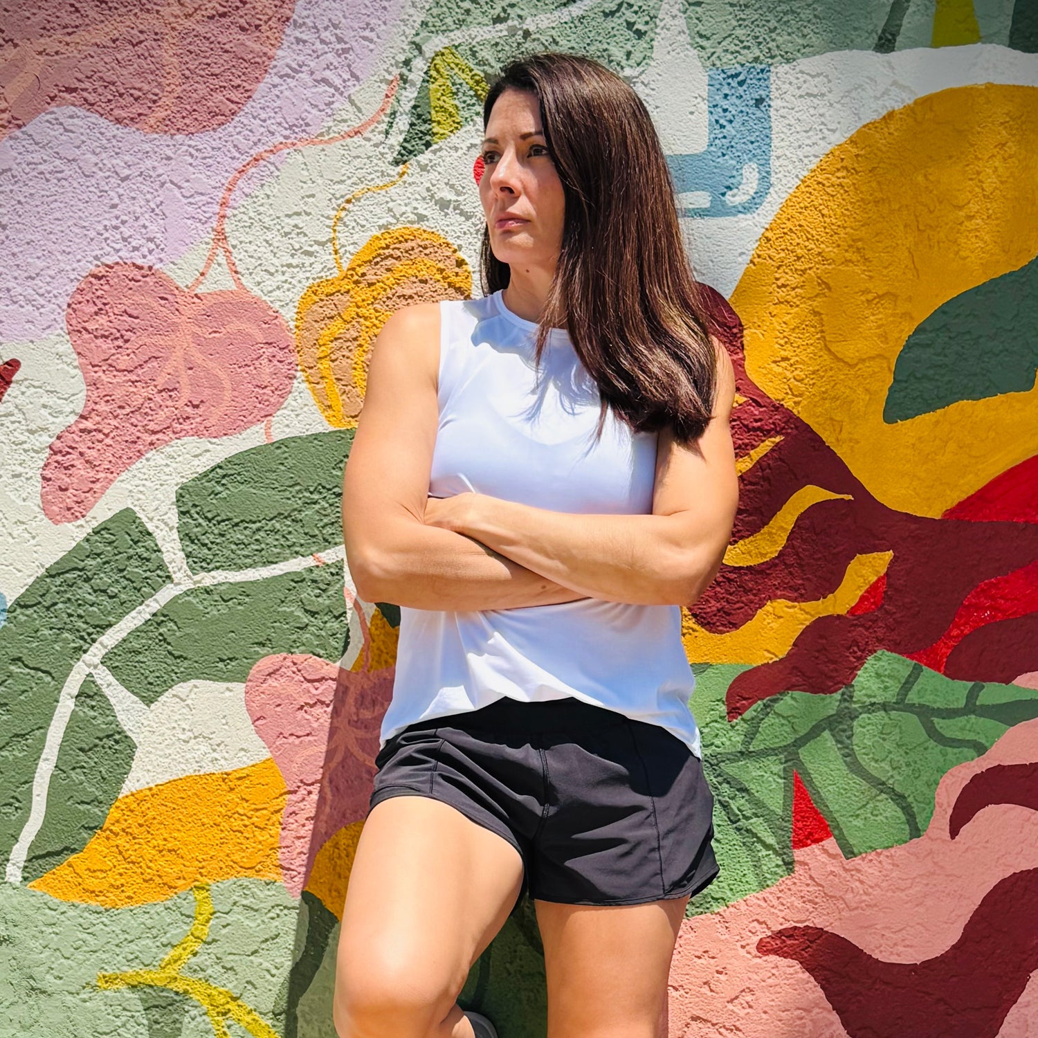 Woman in white tank top and black shorts stands with arms crossed against a colorful mural wall.