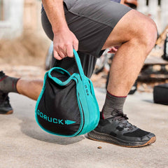While outdoors, a person does a lunge with a teal and black Sand Kettlebells from GORUCK, featuring heavy-duty padded handles.