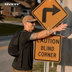 A man wearing a Basic Rucker® weighted rucksack touches a road sign that says "CAUTION BLIND CORNER," with "KEVIN 6’0”" above him.