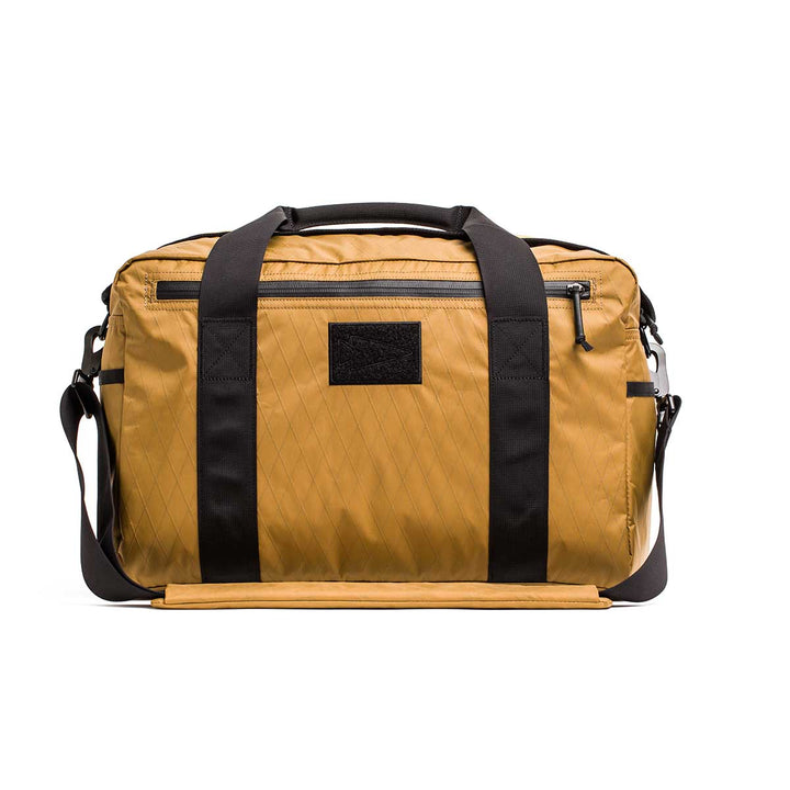 The Kit Bag - X-PAC, inspired by Special Forces gear, features a brown aviator design with black handles, a front zipper pocket, and shoulder strap, shown against a white background.