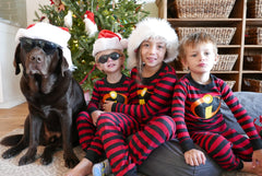Three kids and a dog wear matching “Monster Forever Tee” shirts and Santa hats, sitting by a Christmas tree, celebrating the joy of family—even as they cherish loving memories with their pet memorial ornament under the branches, honoring those waiting at the Rainbow Bridge.