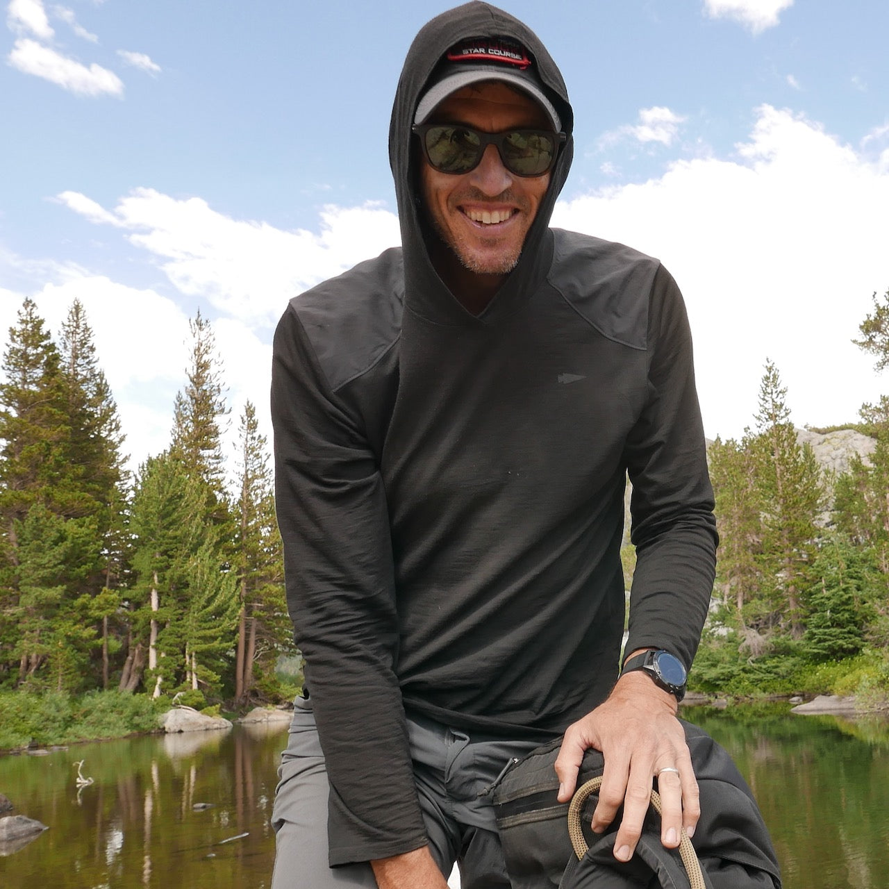 A smiling man in sunglasses enjoys the outdoors near a lake, wearing the Men’s Commando Pullover - Merino Wool with trees in the background.
