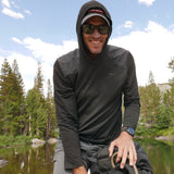 A smiling man in sunglasses enjoys the outdoors near a lake, wearing the Men’s Commando Pullover - Merino Wool with trees in the background.