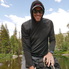 A smiling man in sunglasses enjoys the outdoors near a lake, wearing the Men’s Commando Pullover - Merino Wool with trees in the background.