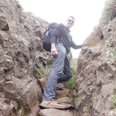 A man with a backpack hikes between rocky cliffs, smiling at the camera on a cloudy day, wearing rugged MACV-2 - Mid Top - Briar + Coyote boots designed for adventure.