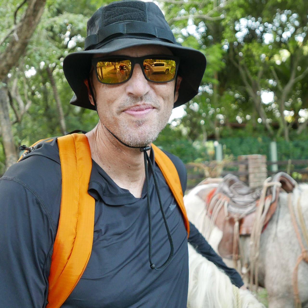Man in sunglasses, a black Boonie Hat - Tactical - ToughDry, and an orange backpack stands outdoors near saddled horses, showcasing a rugged military style.