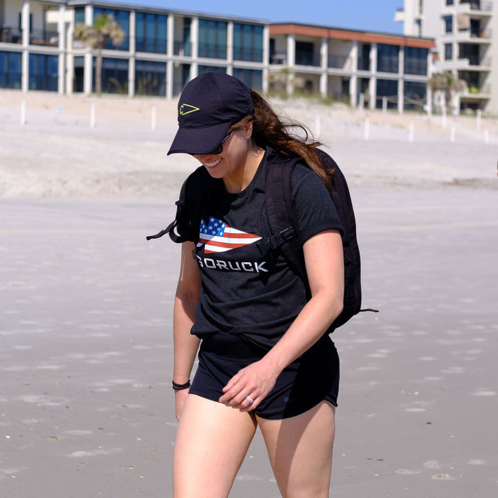 A smiling woman in the GORUCK USA Tee - Tri-Blend and a GORUCK spearhead backpack walks along a sandy beach, with buildings visible in the background.