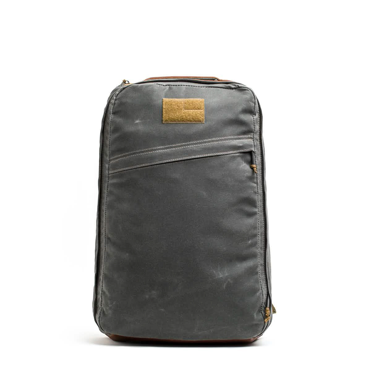 The GR1 USA - Heritage Waxed Canvas backpack in grey with a brown patch and zippers stands upright against a white background.