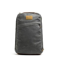The GR1 USA - Heritage Waxed Canvas backpack in grey with a brown patch and zippers stands upright against a white background.