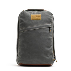 The GR1 USA - Heritage Waxed Canvas is a gray backpack featuring a diagonal zipper pocket and a tan front patch, crafted for timeless style.