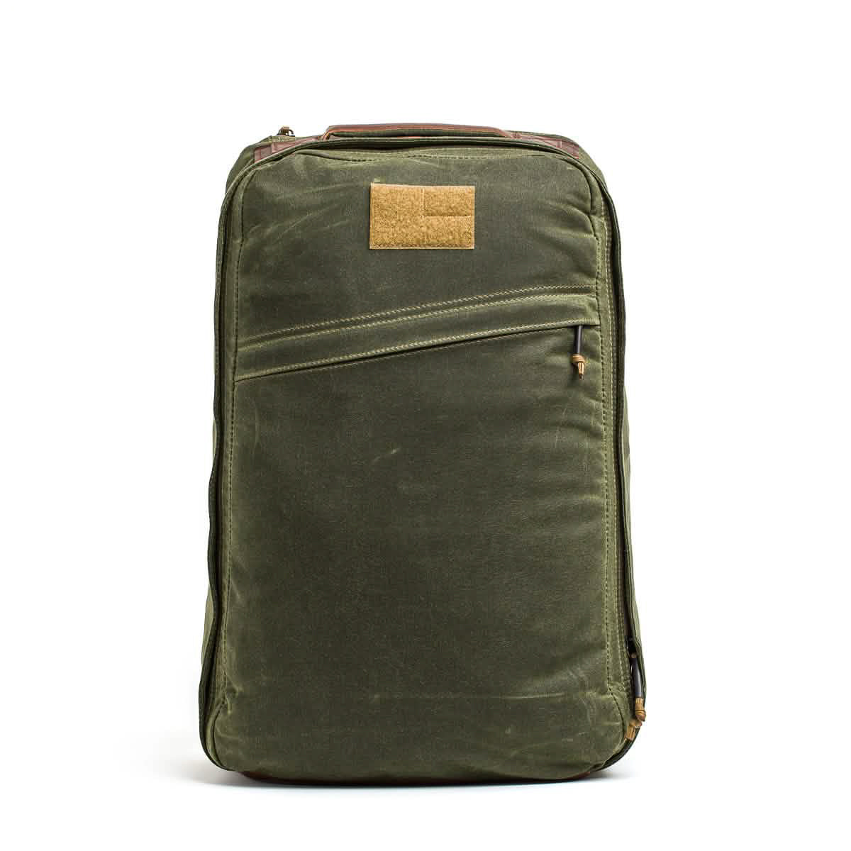 The GR1 USA - Heritage Waxed Canvas is an olive green retro canvas backpack featuring a front zipper pocket and a tan rectangular patch near the top, combining classic style with everyday functionality.