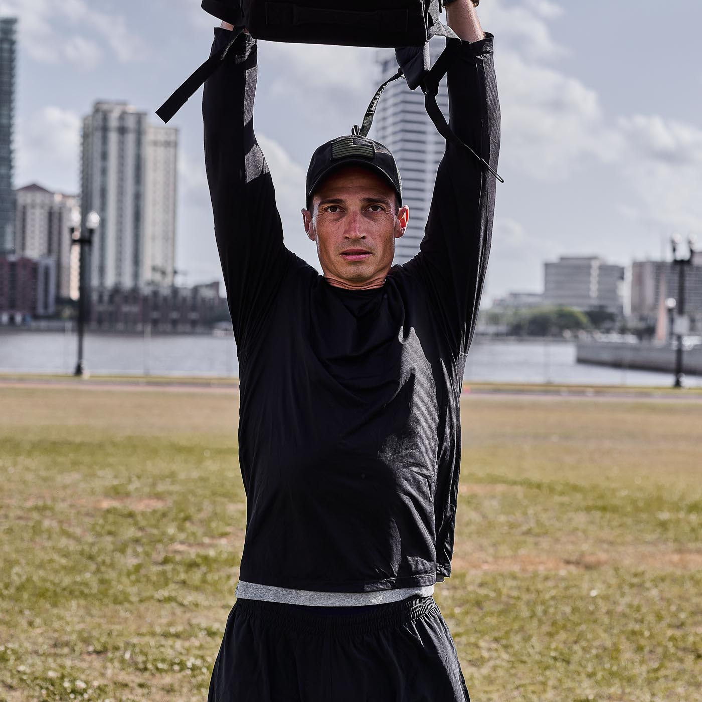 A man in black Men's USA Long Sleeve Performance Tee - ToughMesh lifts a weighted bag overhead outdoors, city buildings behind him, demonstrating the tee's durable ToughMesh™ fabric.