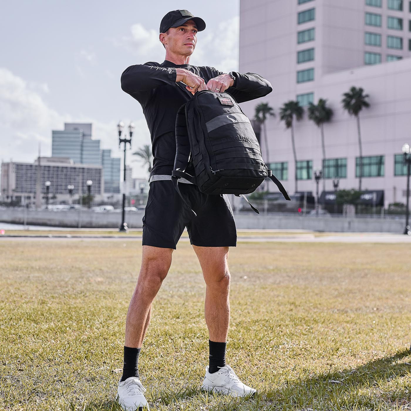 A man in athletic wear stands on grass with city buildings and palm trees behind him, wearing the Men's USA Long Sleeve Performance Tee made from ToughMesh™ fabric and holding a black backpack.