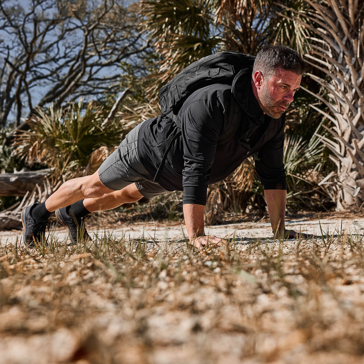 A man does push-ups outdoors on rocky ground in the Italian Alps, wearing the Men's USA Long Sleeve Performance Full Zip Hoodie - ToughMesh, athletic clothing, a backpack, and surrounded by trees.