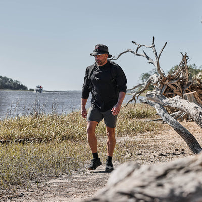 A man wearing the Men's USA Long Sleeve Performance Full Zip Hoodie - ToughMesh, a hat, and a backpack walks along a sandy riverside trail with driftwood and tall grass.