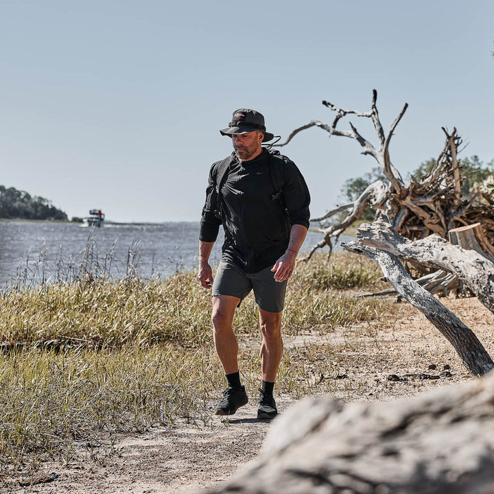 A man wearing the Men's USA Long Sleeve Performance Full Zip Hoodie - ToughMesh, a hat, and a backpack walks along a sandy riverside trail with driftwood and tall grass.