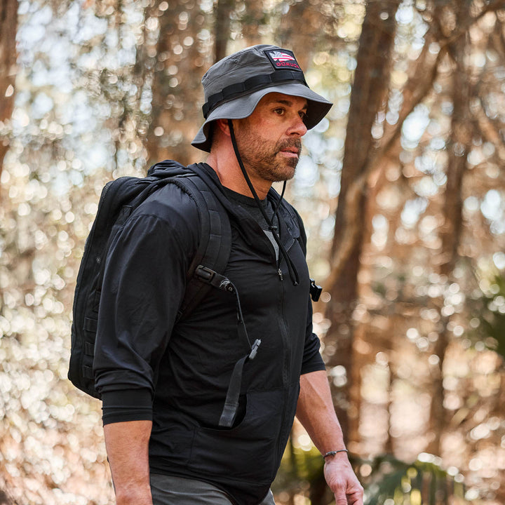 A man wearing the Men's USA Long Sleeve Performance Full Zip Hoodie - ToughMesh and a black bucket hat walks through a sunlit forest carrying a backpack.