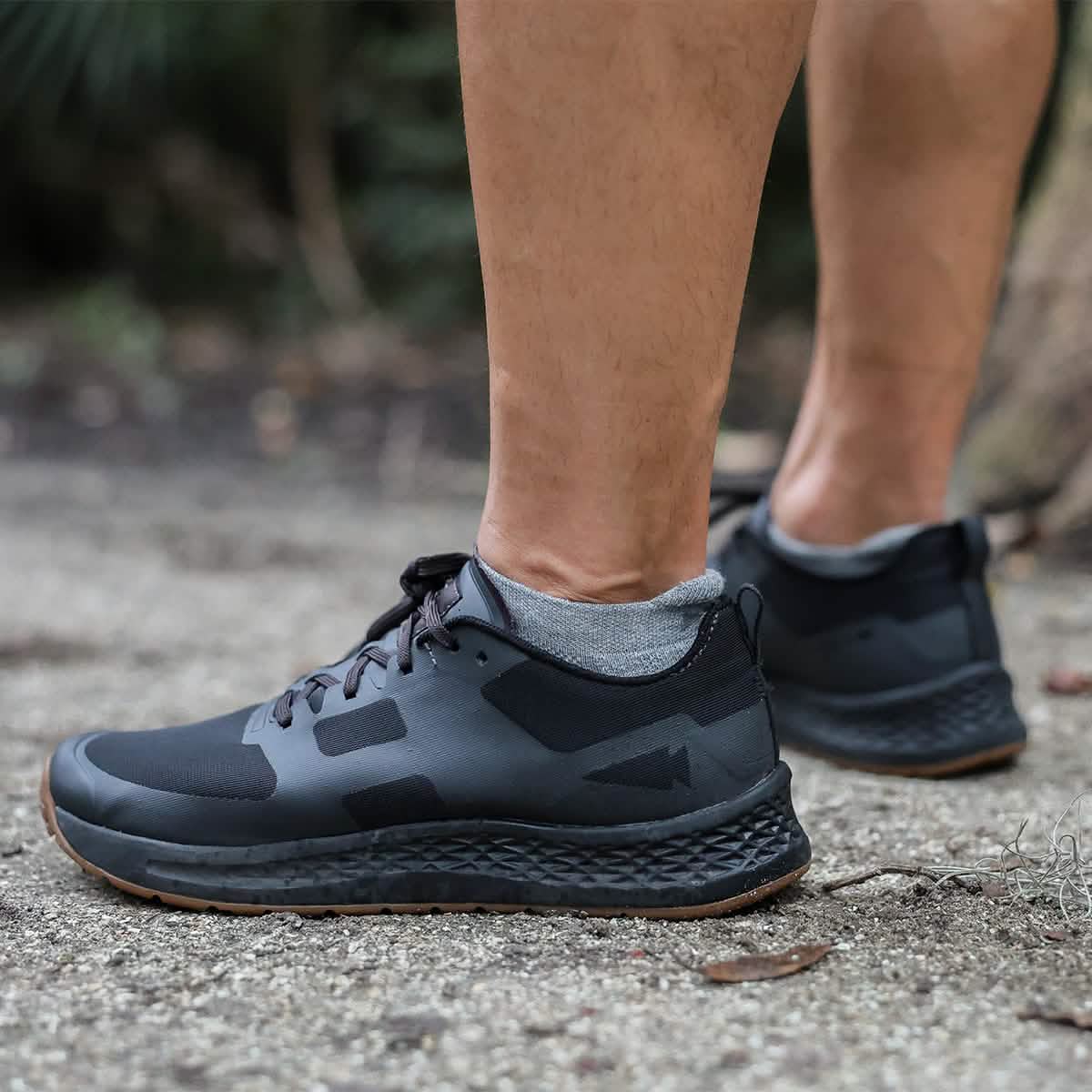 A person wearing black athletic shoes and gray GORUCK Merino Challenge Socks - Ankle stands on a gravel path. The focus is on the shoes, highlighting their design and texture, while the comfort of these high-quality socks made in the USA enhances every step.