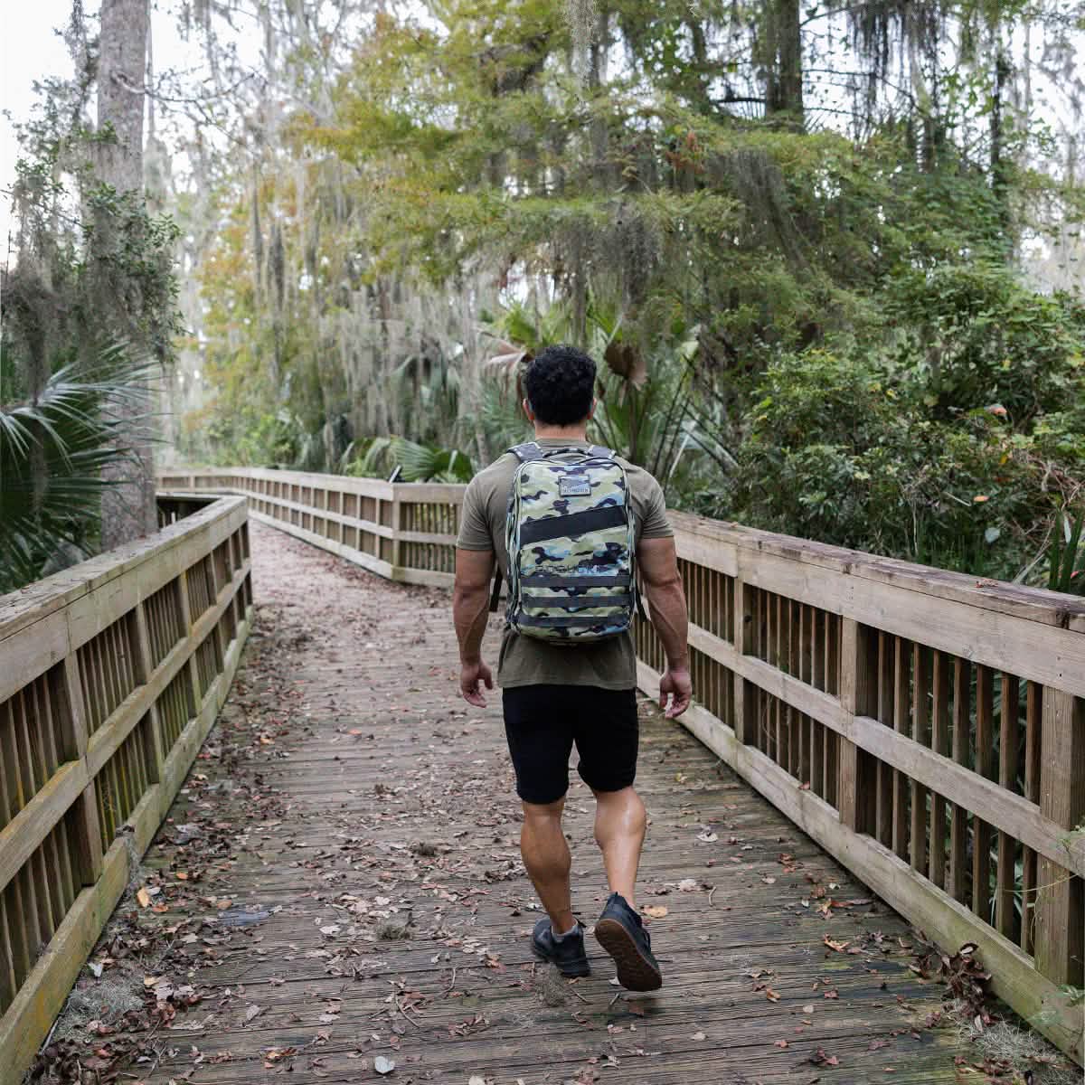 A person sporting a camo backpack walks along a wooden boardwalk through a forested area, surrounded by lush green foliage and scattered leaves. Dressed in a green shirt, black shorts, and GORUCK Merino Challenge Socks - Ankle, they enjoy the tranquility of nature.