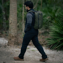 Man hiking in forest wearing black and gray outfit with olive green GORUCK backpack and brown boots