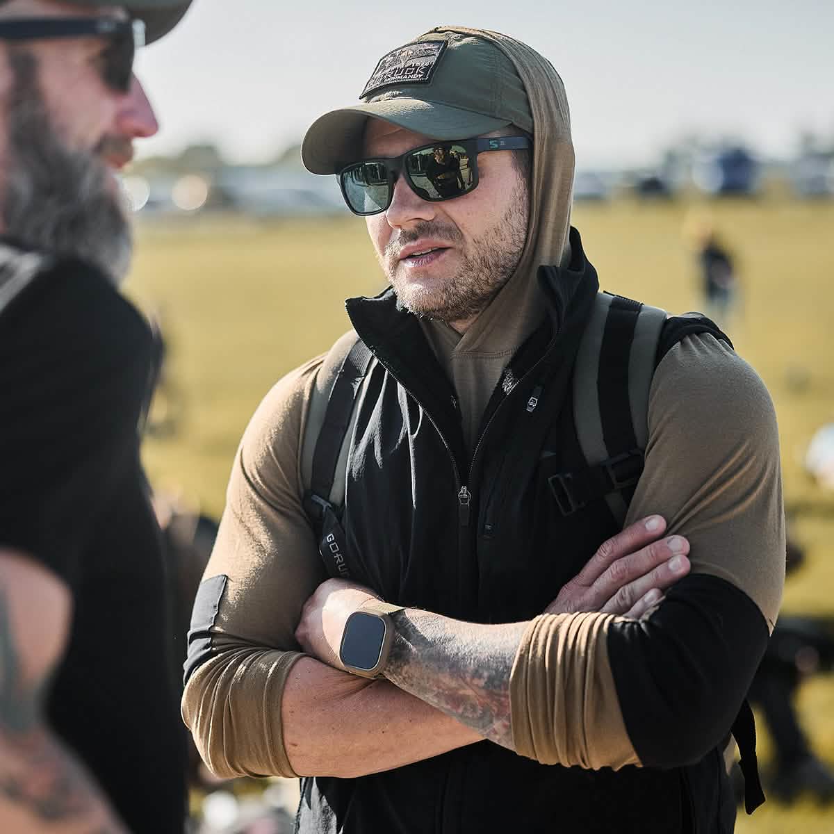 A man wearing a pair of sunglasses, a cap, and the Men’s Commando Pullover - Merino Wool from GORUCK stands outdoors with his arms crossed. He is talking to another person with a beard. The background features an open field and blurred figures.